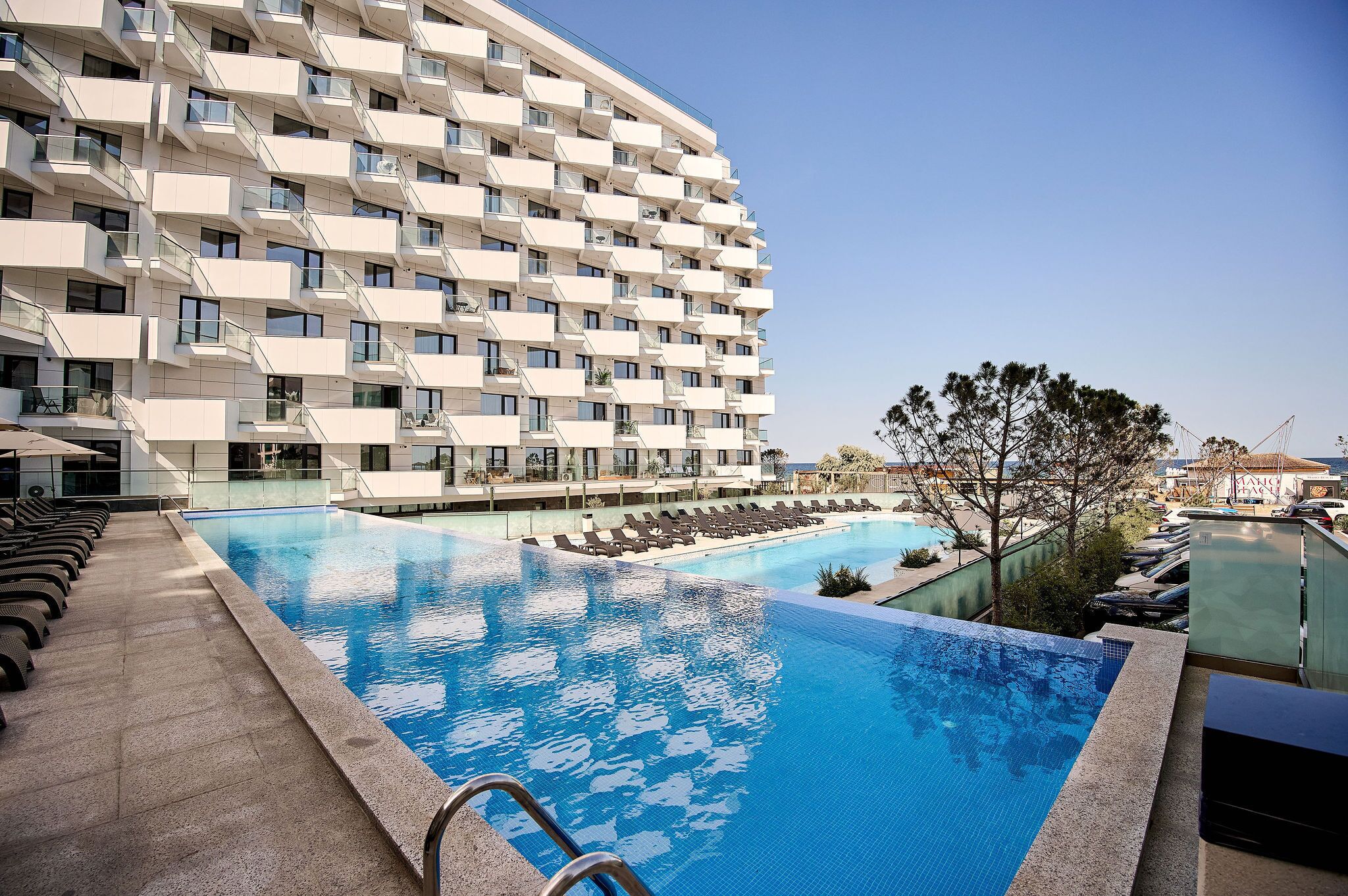 A modern hotel with a large swimming pool, multiple balconies, and a clear view of the surrounding area.