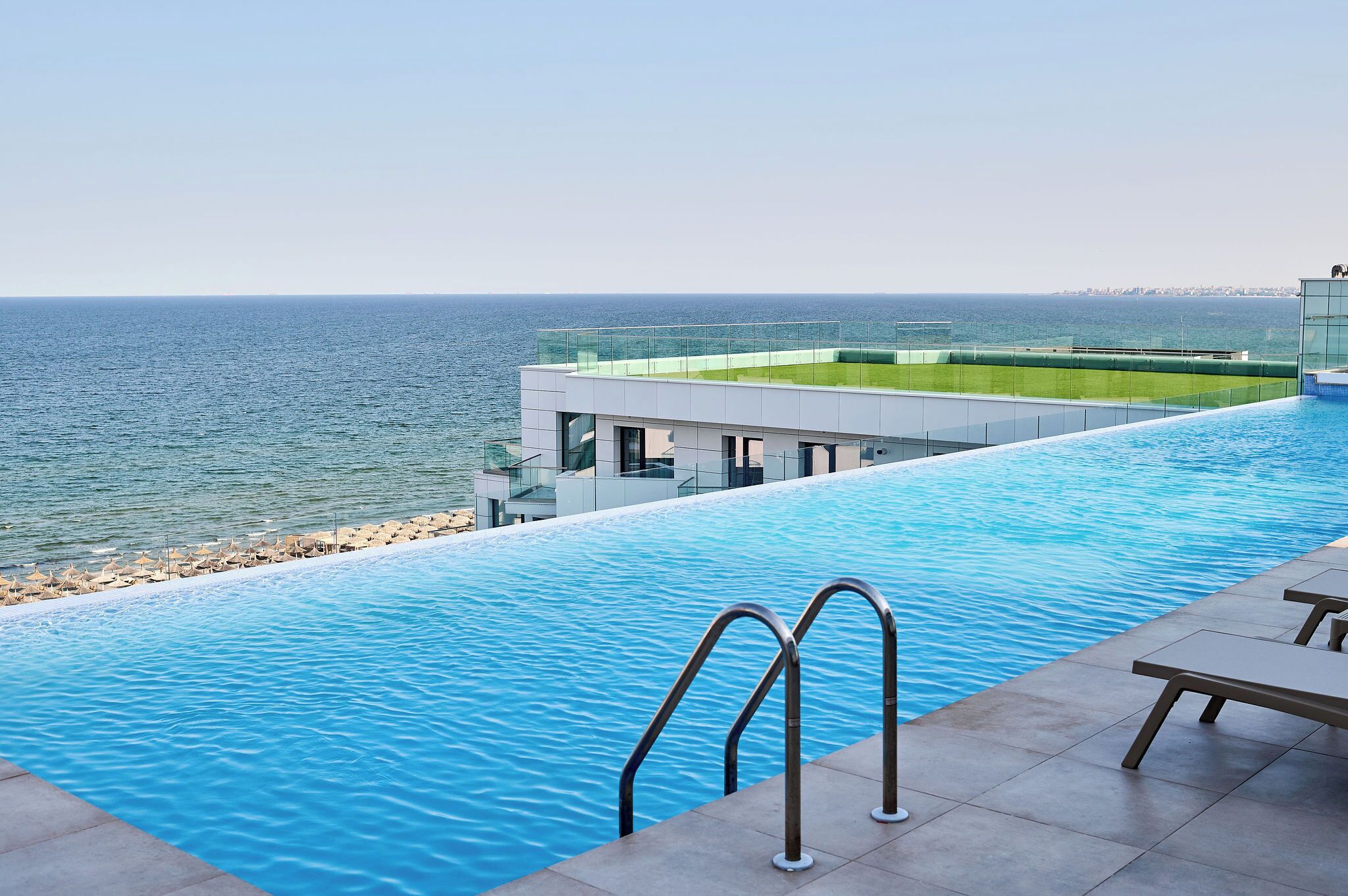 A modern infinity pool with a clear view of the sea, a glass balustrade, and a section of the poolside area with a lounge chair.