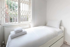 A single bed with white bedding and a white pillow in a room with a white radiator and a window with a white lattice design. - Spacious & Welcoming 4BD House - Kennington (London)