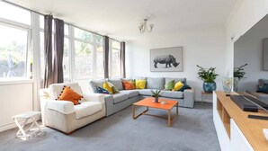 Ein modernes Wohnzimmer mit einem grauen Ecksofa, einem weißen Sessel und einem Holztisch. Ein großes Fenster mit Vorhängen, eine Topfpflanze und ein gerahmtes Tierbild an der Wand sind zu sehen.