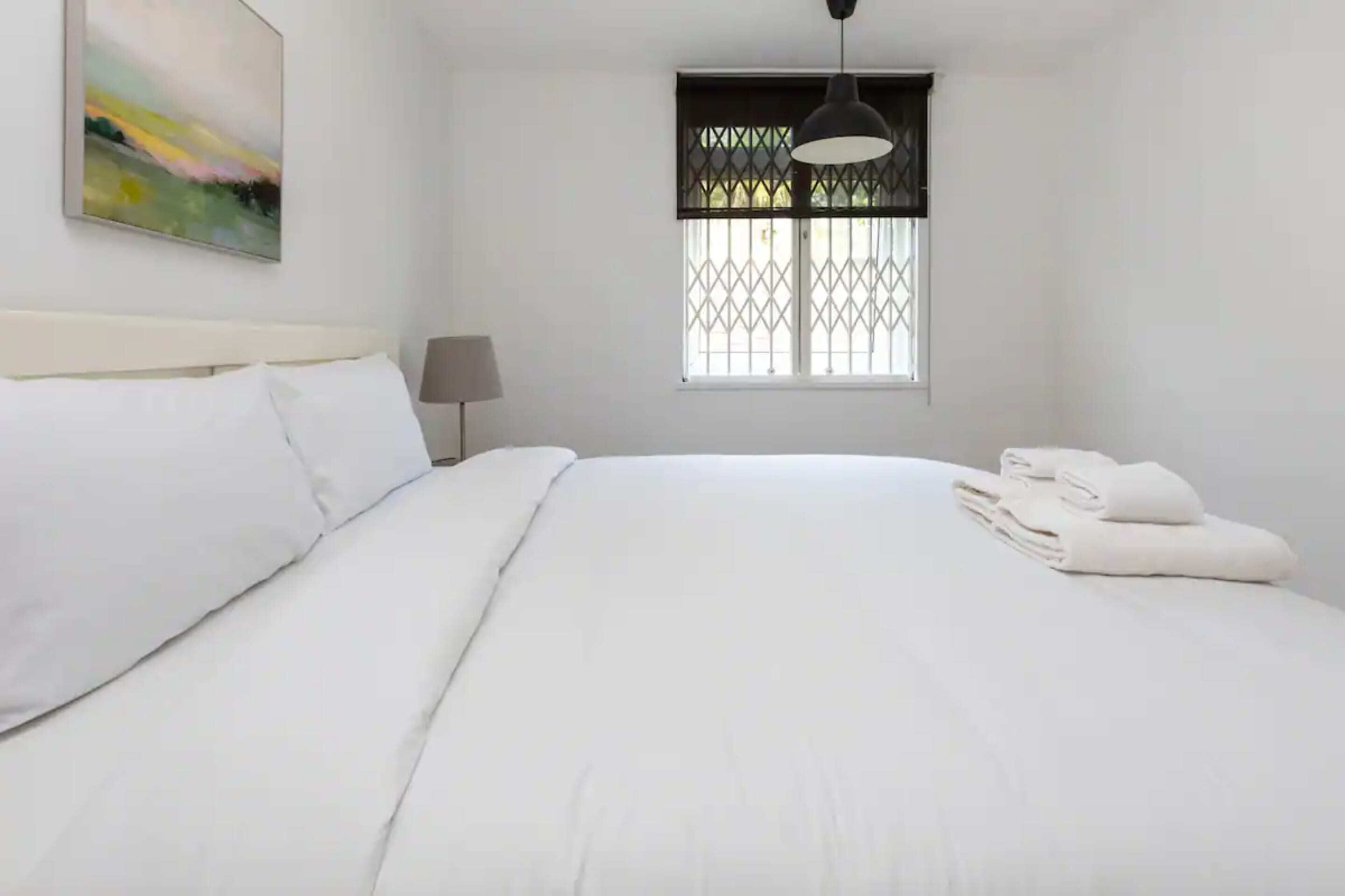 Un lit bien fait, avec du linge de lit blanc, une lampe de chevet et une fenêtre ornée d’une grille en métal.