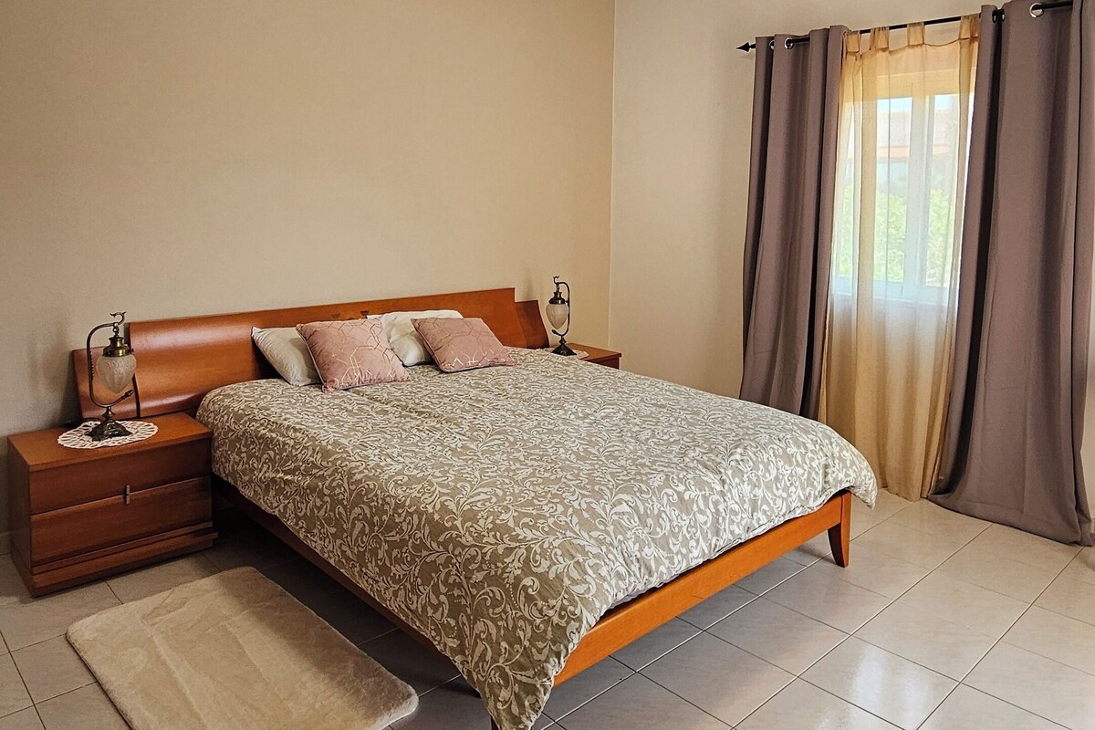 Une chambre à coucher avec un lit en bois, une table de chevet, une lampe de chevet, un lampadaire, une fenêtre avec des rideaux et un luminaire au plafond.