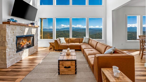 A modern living room with a stone fireplace, leather sofa, wooden coffee table, and large windows offering a mountain view. - Elegant Cabin | Indoor Pool, Dog-Friendly (Sevierville)