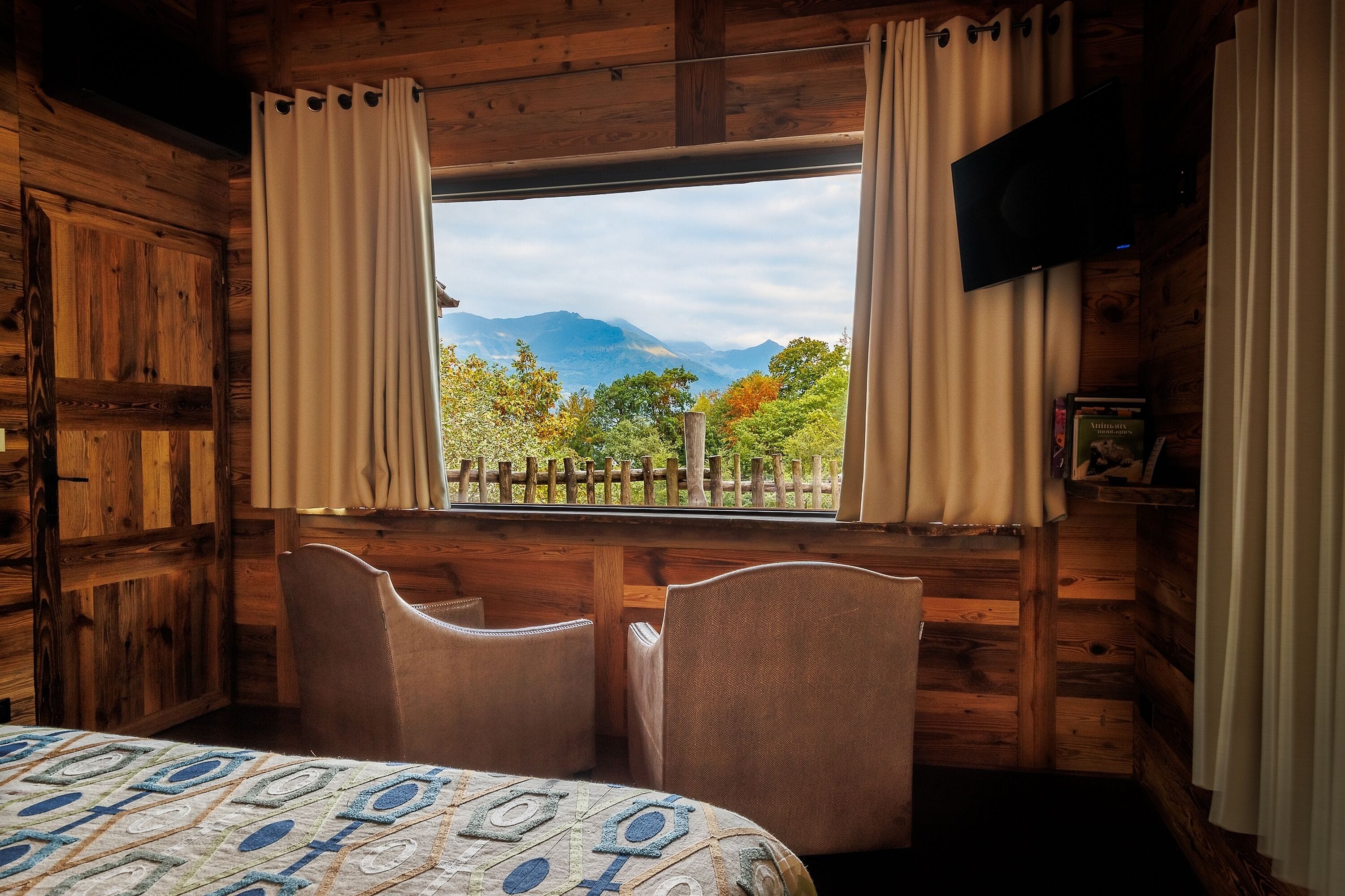 A cozy cabin room with a large window, wooden walls, a bed, two chairs, and a mounted TV.