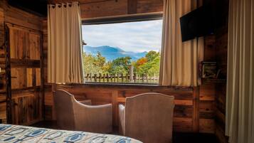 A cozy cabin room with a large window, wooden walls, a bed, two chairs, and a mounted TV.