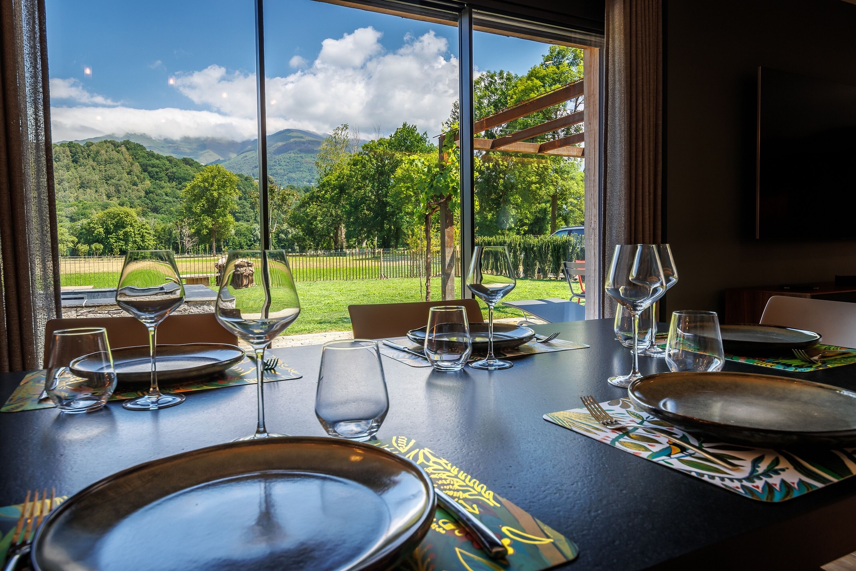 Una mesa de comedor dispuesta con platos, vasos y cubiertos, con vistas a un paisaje exuberante con montañas y una valla.