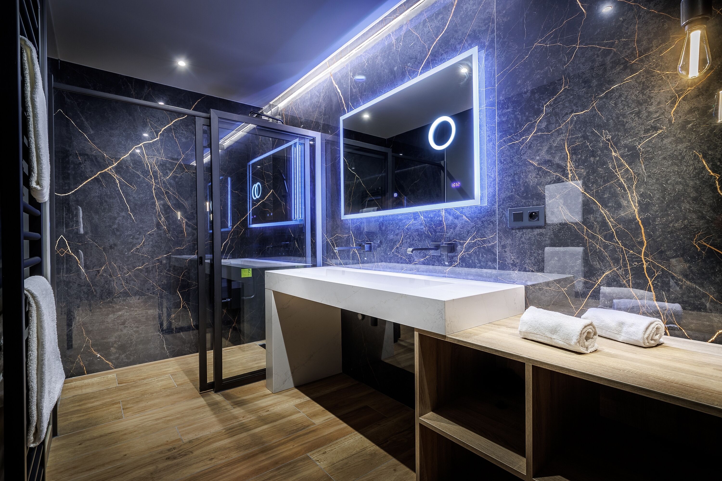 A modern bathroom with a marble backsplash, a large mirror with integrated lighting, and a wooden vanity.