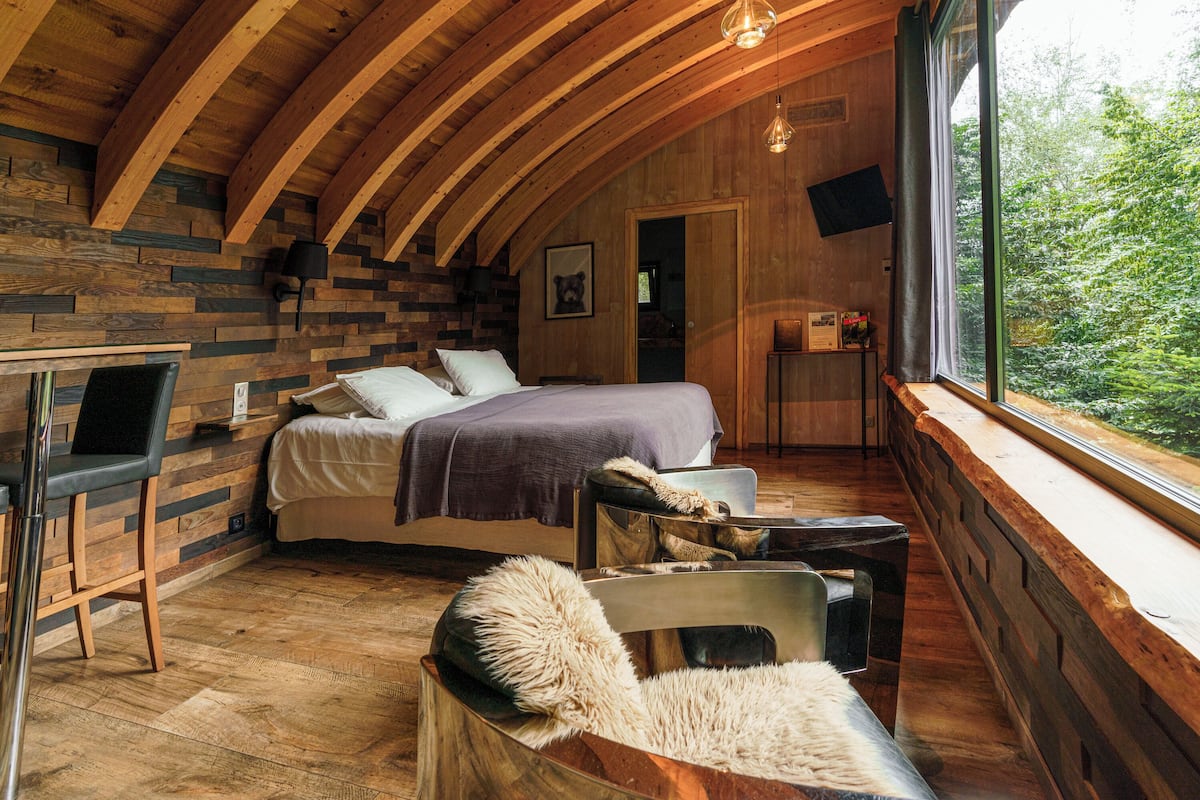 A cozy bedroom with a wooden ceiling, a bed, a desk, and a large window overlooking greenery.