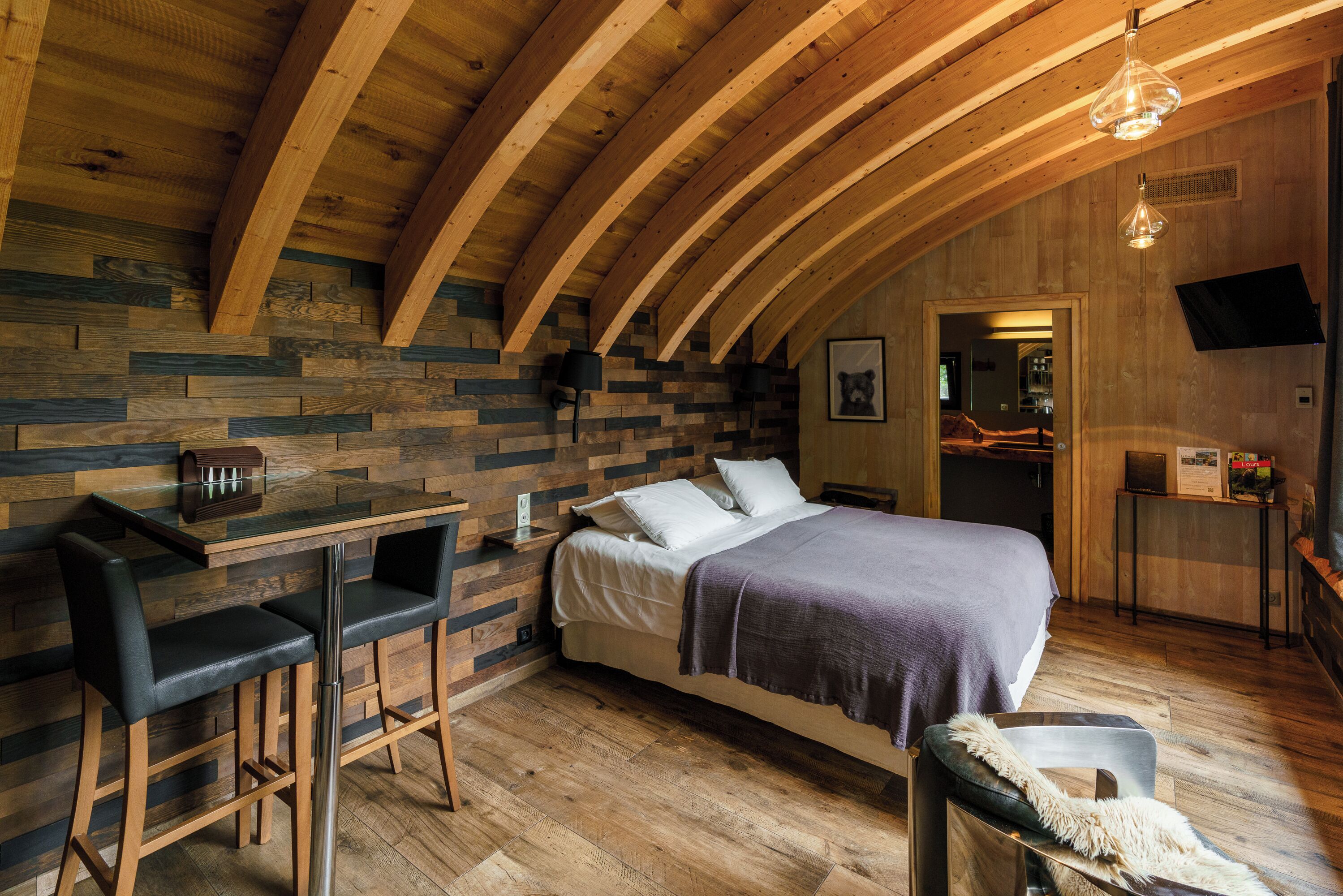 A cozy room with a wooden ceiling, a bed with a purple blanket, a small table with chairs, and a TV mounted on the wall.