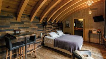 A cozy room with a wooden ceiling, a bed with a purple blanket, a small table with chairs, and a TV mounted on the wall.