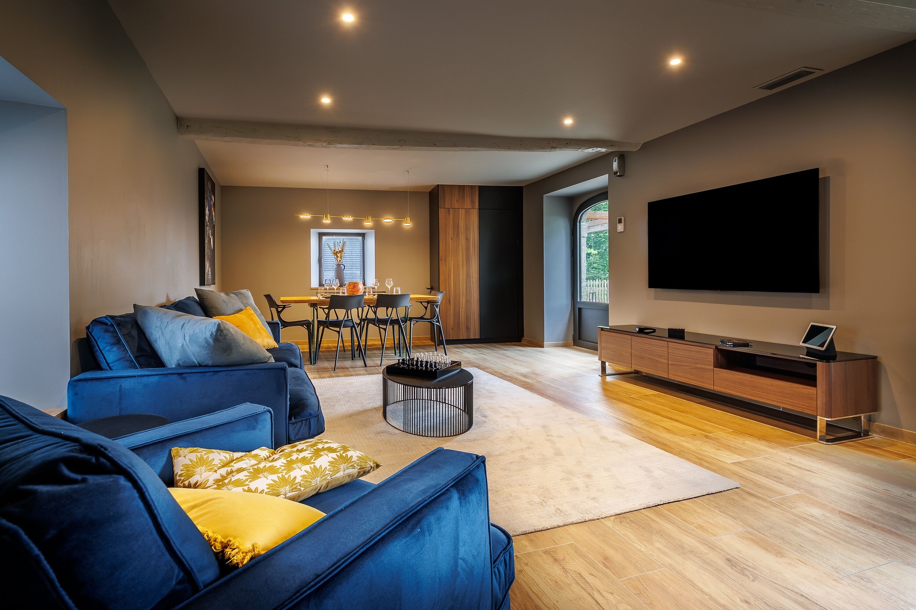 A modern living room with a blue sofa, a dining area with a table and chairs, and a large flat-screen TV mounted on the wall.