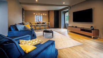 A modern living room with a blue sofa, a dining area with a table and chairs, and a large flat-screen TV mounted on the wall.