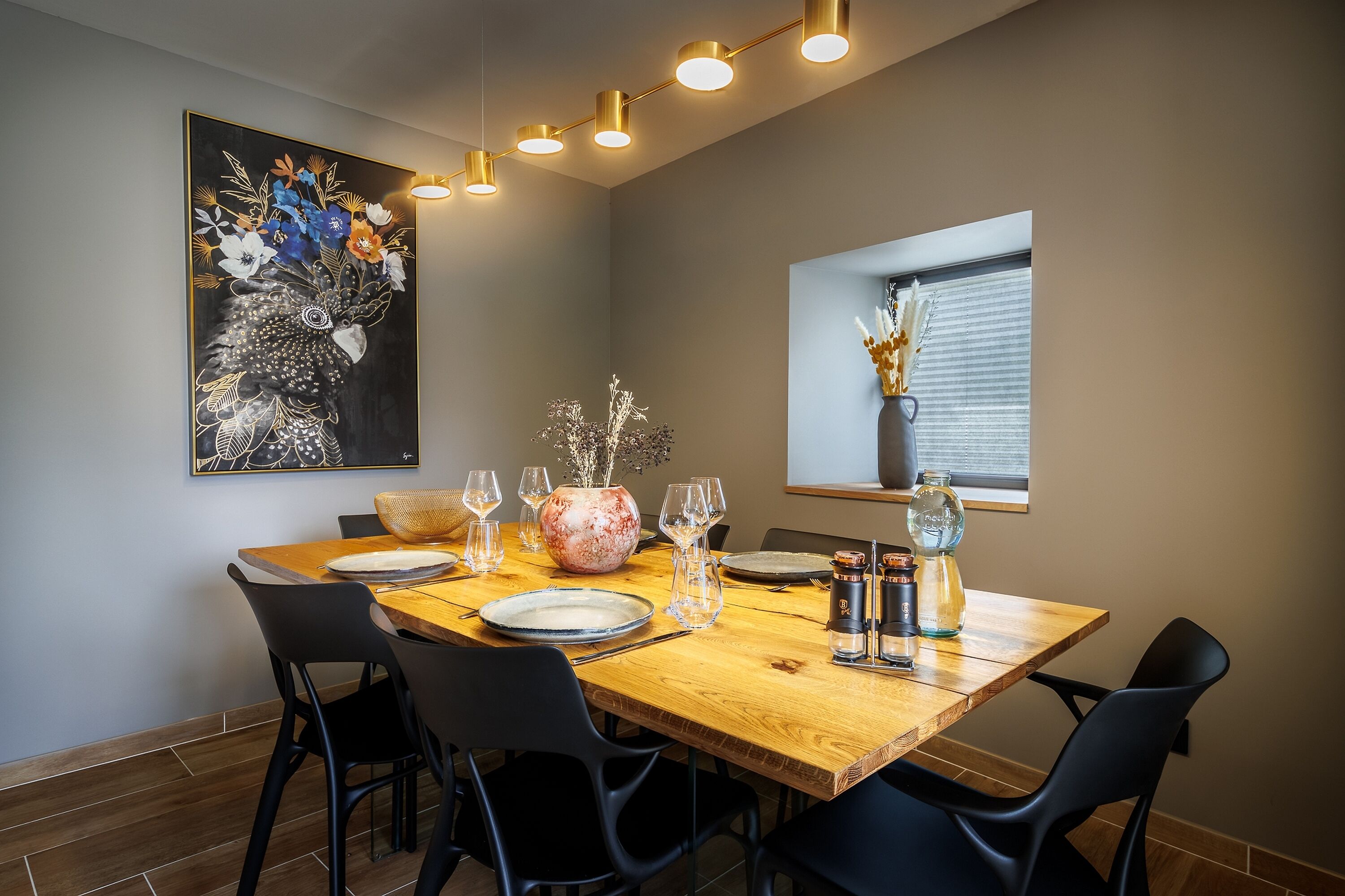 Un comedor con una mesa de madera, sillas negras, un cuadro floral grande y un florero con flores.