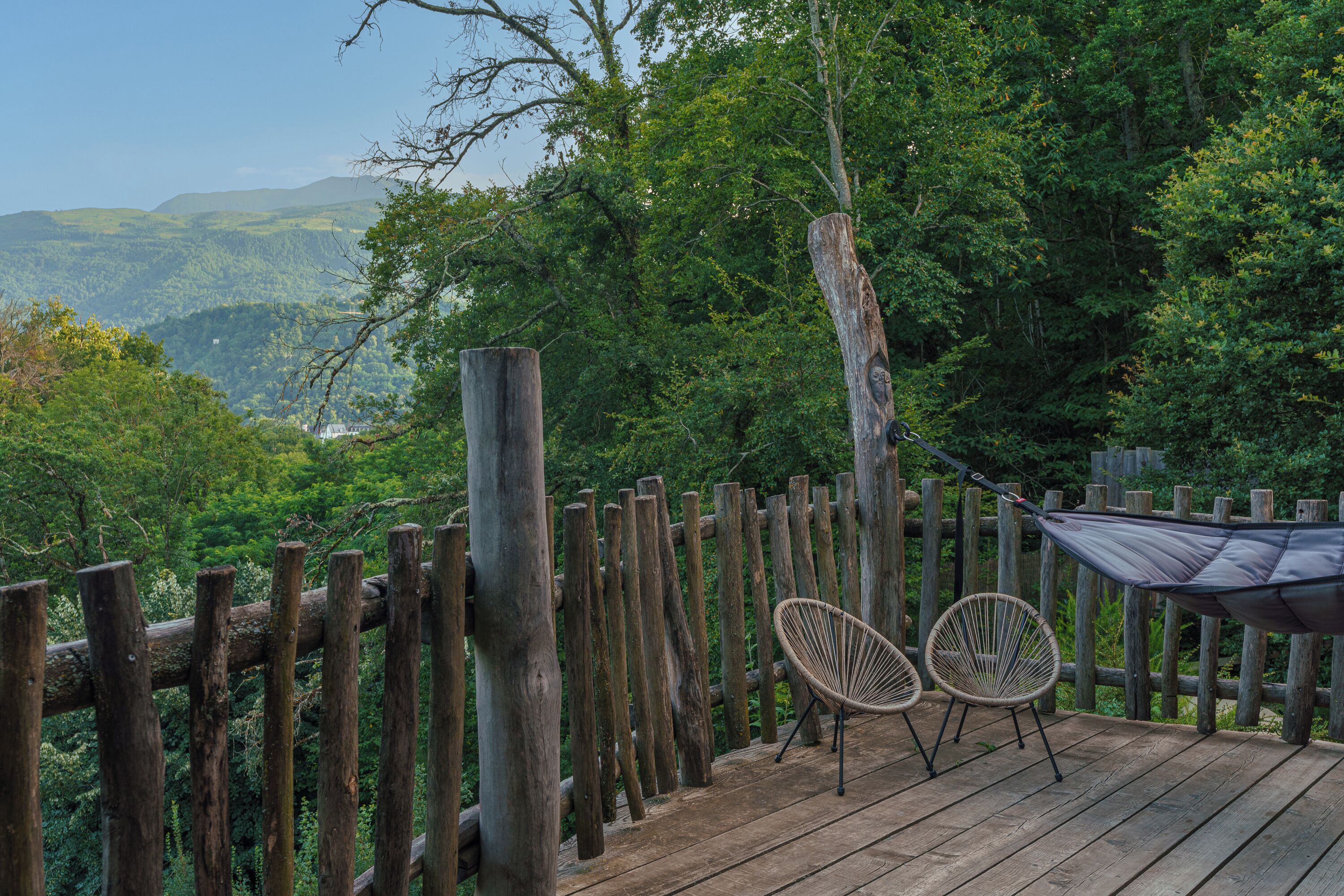 Une terrasse en bois avec deux fauteuils en osier et un hamac, d’où l’on peut admirer un paysage boisé.