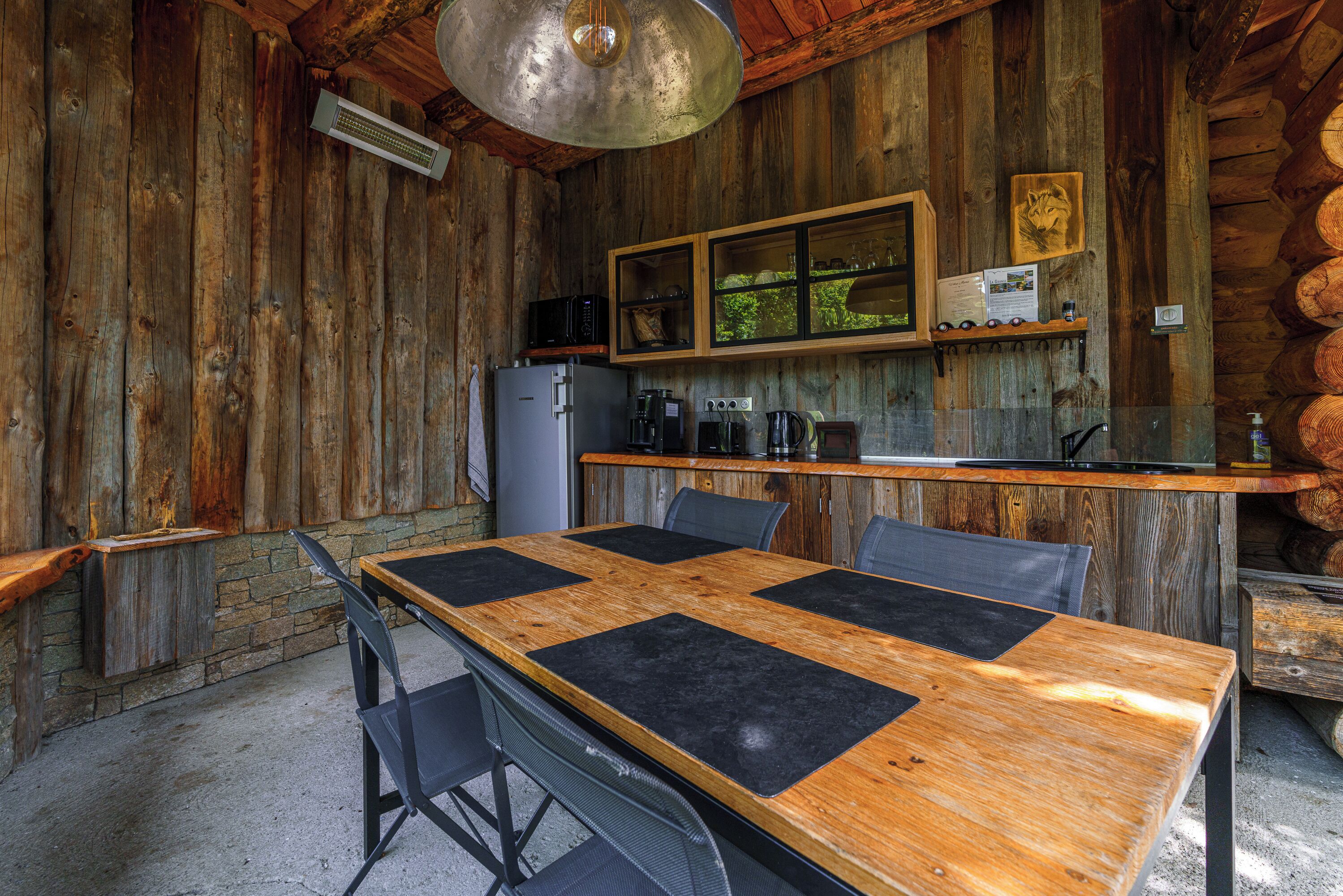 A rustic wooden interior with a large wooden table, chairs, a refrigerator, a coffee maker, and a wooden shelf with various items.