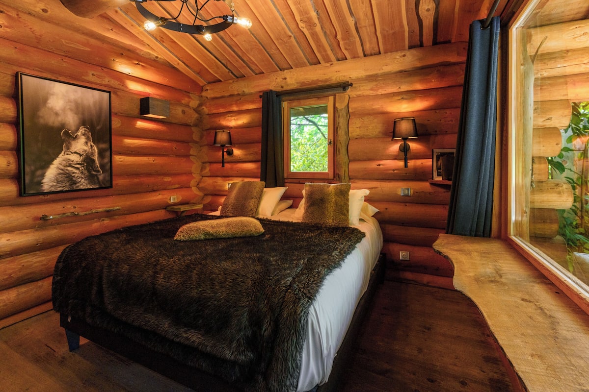 A log cabin bedroom with a large bed, two wall-mounted lamps, a framed picture, and a window with curtains.
