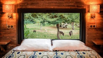 A bed with a patterned bedspread and pillows, a television screen displaying a view of a grassy area with trees and dogs, and wooden walls with wall-mounted lights.