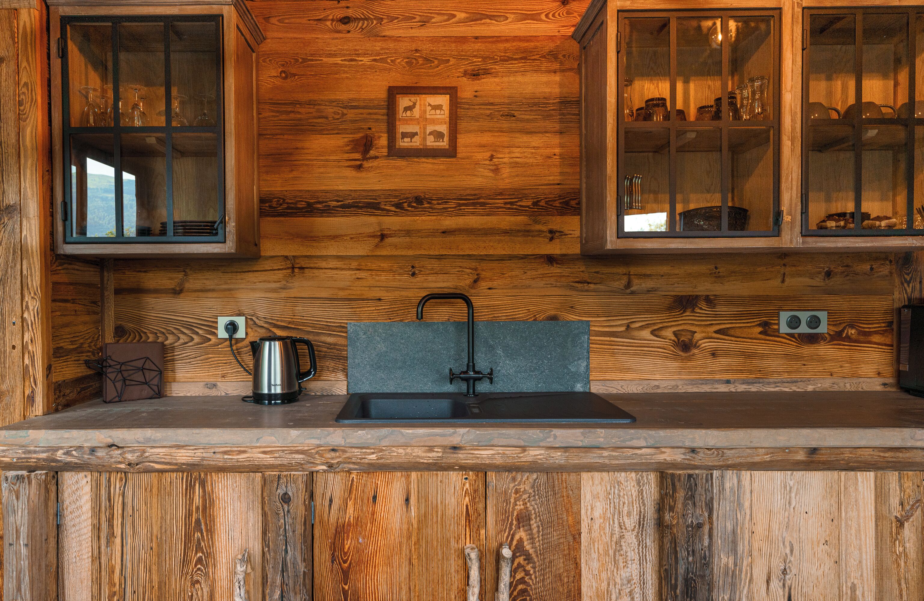 A wooden cabin interior with a kitchen area featuring a sink, a kettle, and cabinets with glass doors.