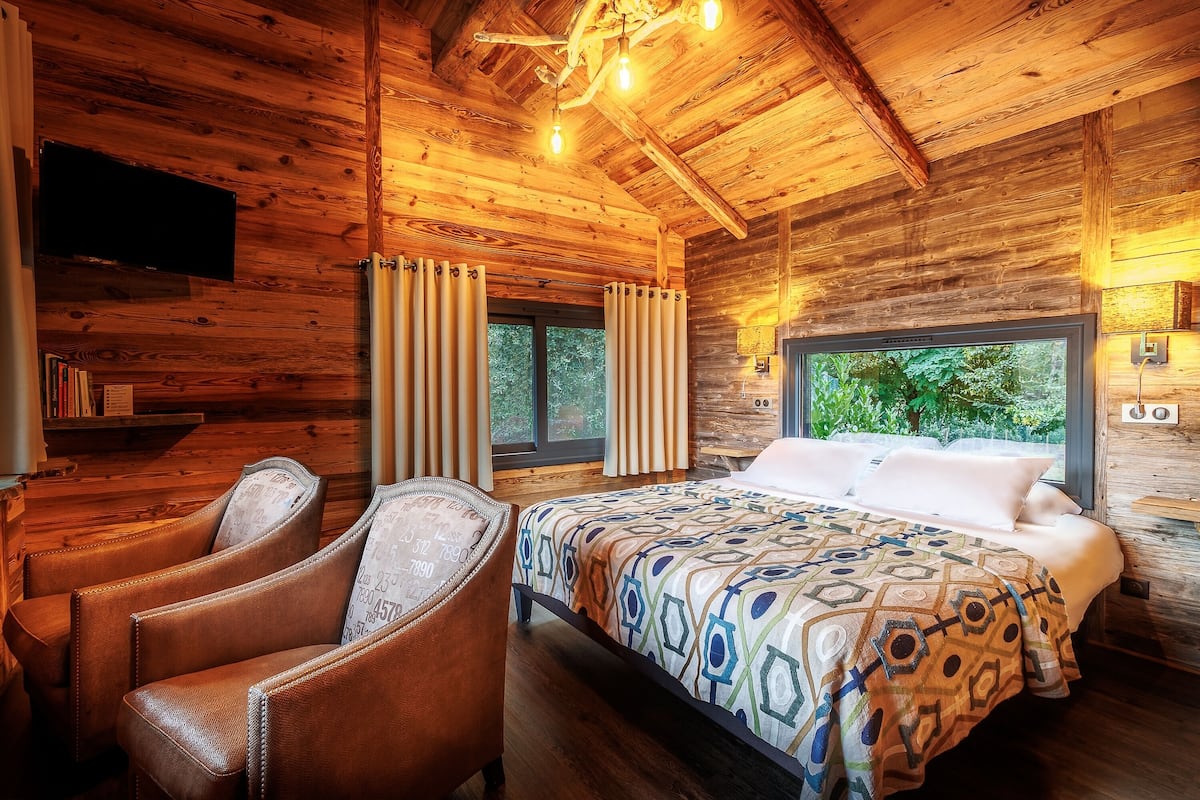 A wooden cabin room with a bed, two leather chairs, a TV, and a window with curtains.