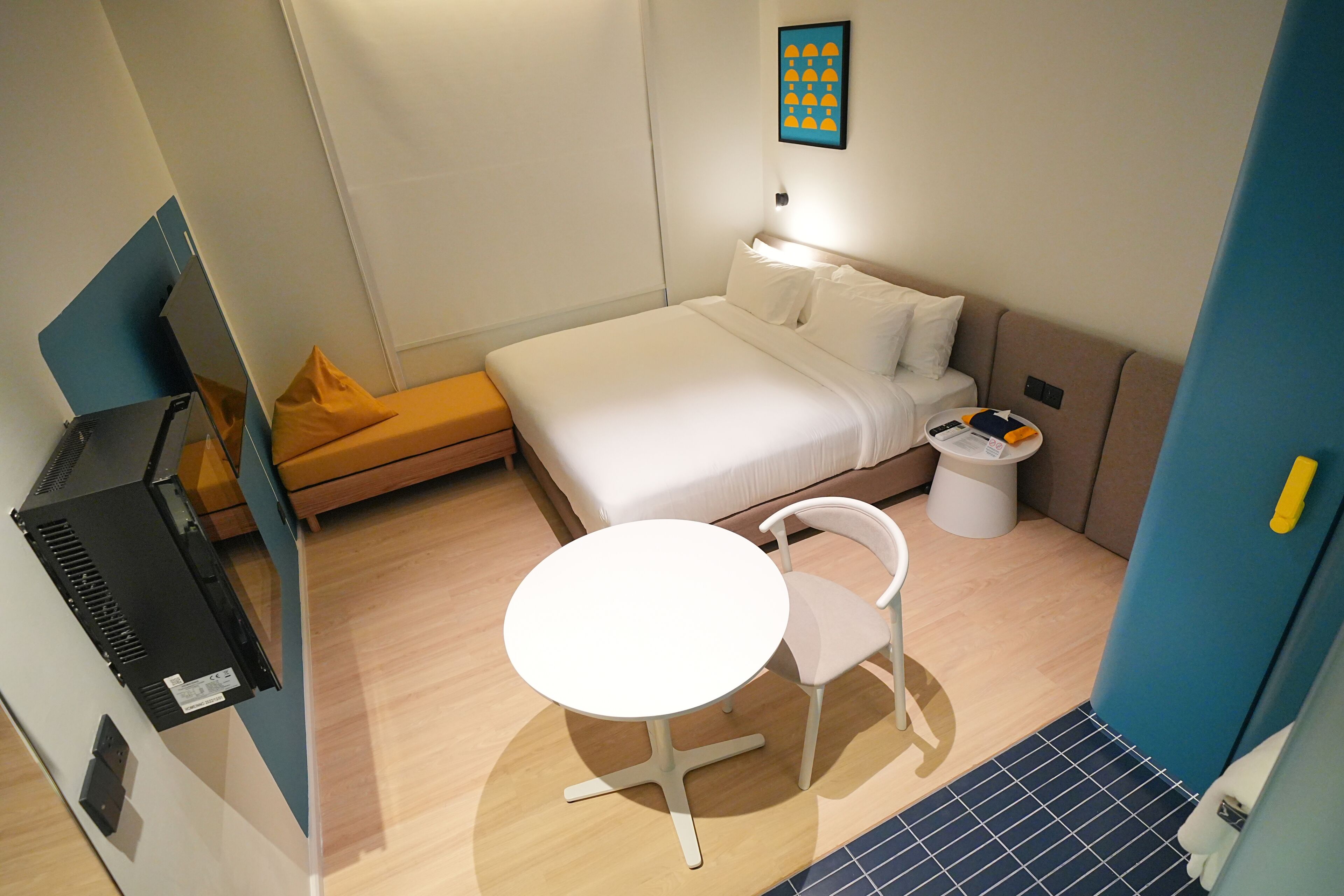 A compact hotel room with a bed, a small table, a chair, a bedside table, and a wall-mounted artwork.