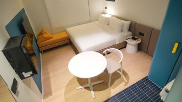 A compact hotel room with a bed, a small table, a chair, a bedside table, and a wall-mounted artwork.