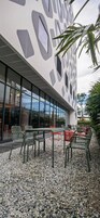 An outdoor seating area with chairs and tables, surrounded by pebbles, in front of a building with a unique geometric facade and large windows.