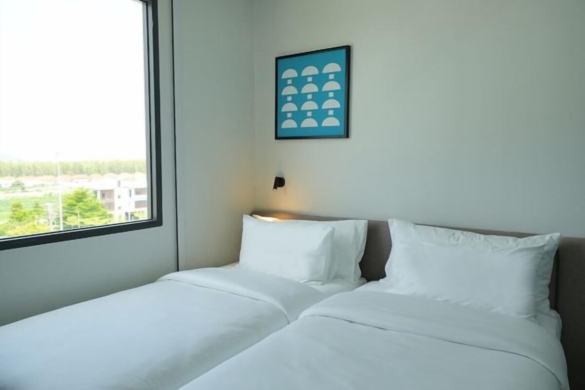 Une chambre d’hôtel avec un lit, une fenêtre donnant sur des immeubles et un tableau encadré au mur.