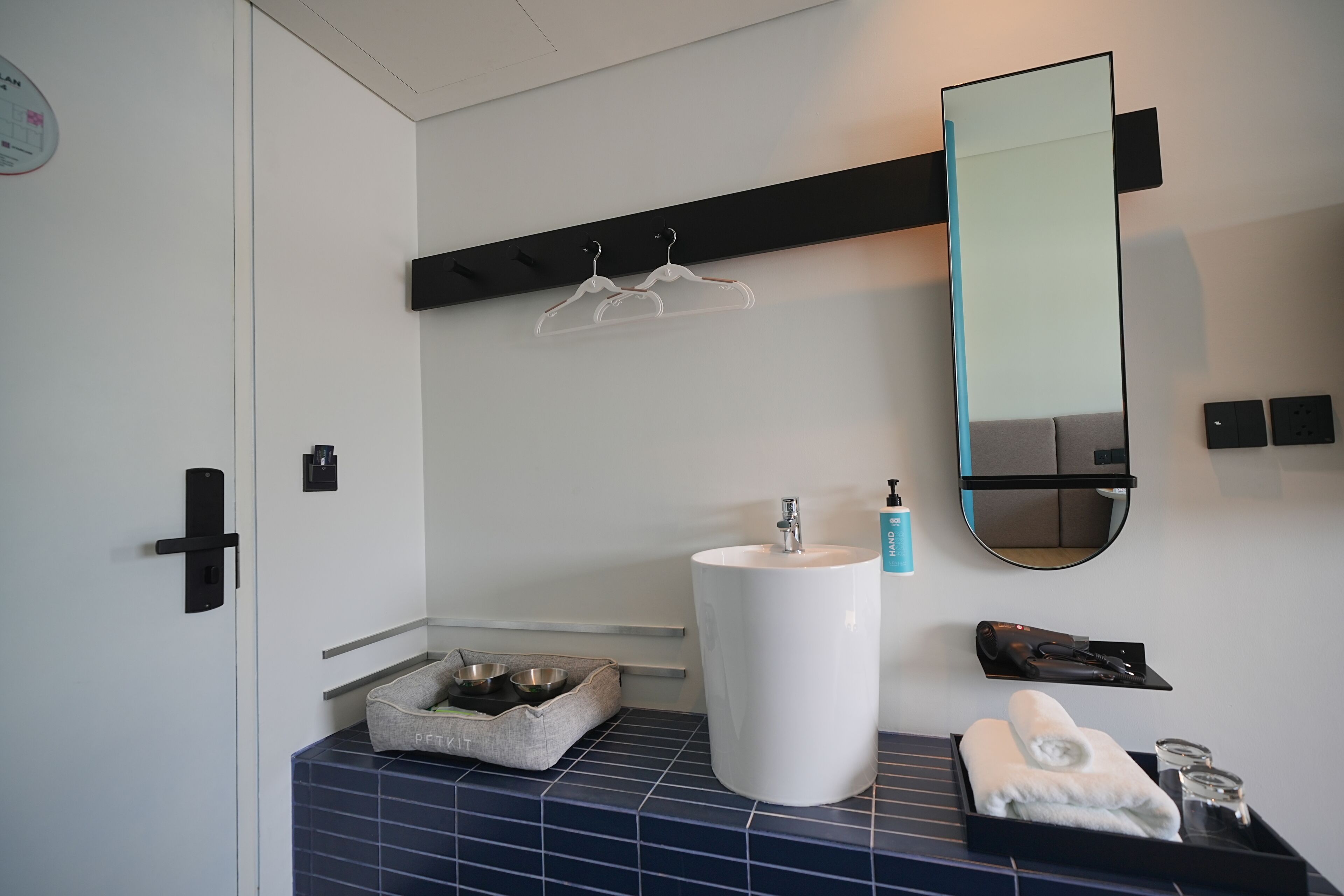 A modern bathroom with a white sink, a mirror, a towel rack, and a pet bed.