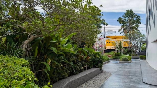 A modern building with a curved facade, a yellow structure in the background, and a pathway lined with greenery. - GO Hotel Chonburi at Central Chonburi (Chonburi)