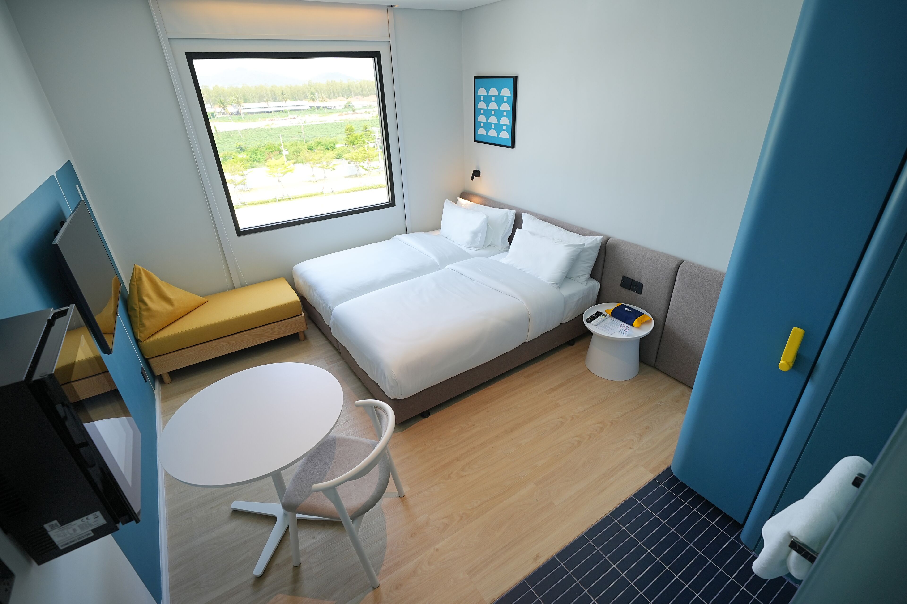 A modern hotel room with a bed, a small table, a chair, a window with a view, and a door.