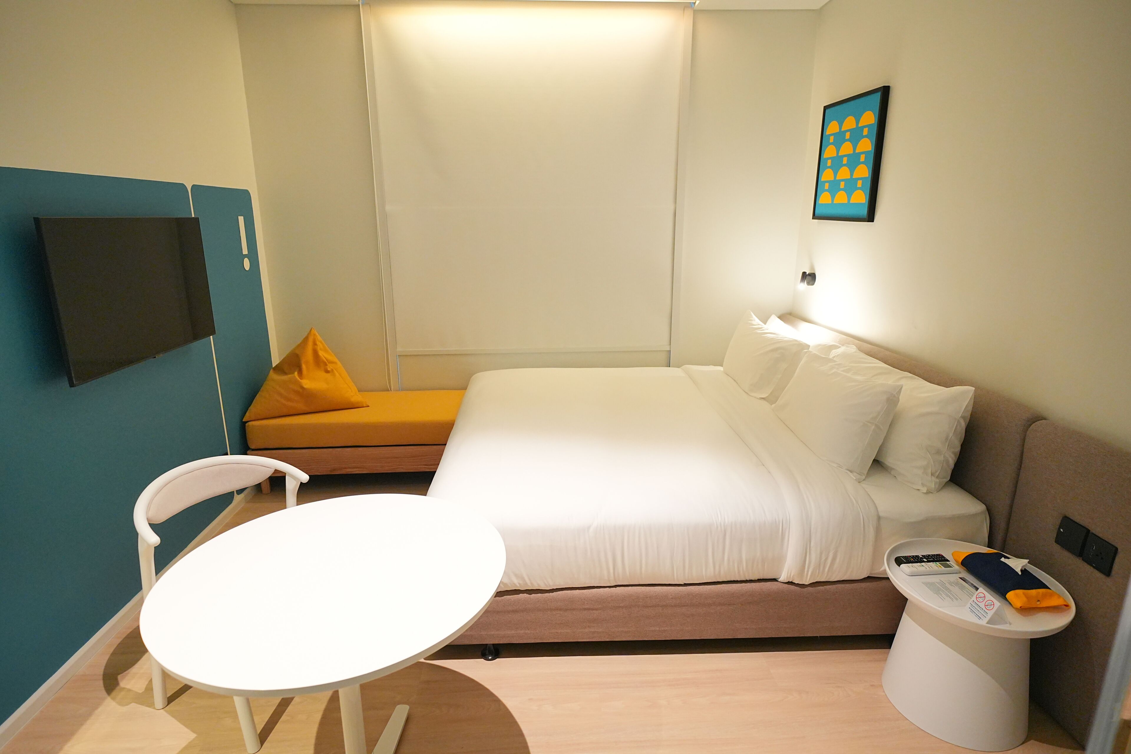 A hotel room with a bed, a small table, a chair, a bedside table with a remote control, and a wall-mounted TV.