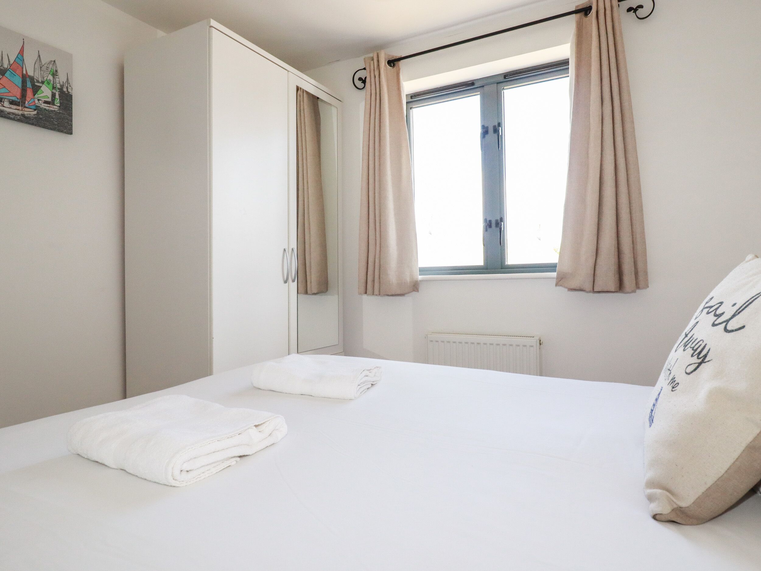 A neatly made bed with a pillow, a window with curtains, a radiator, and a wardrobe.