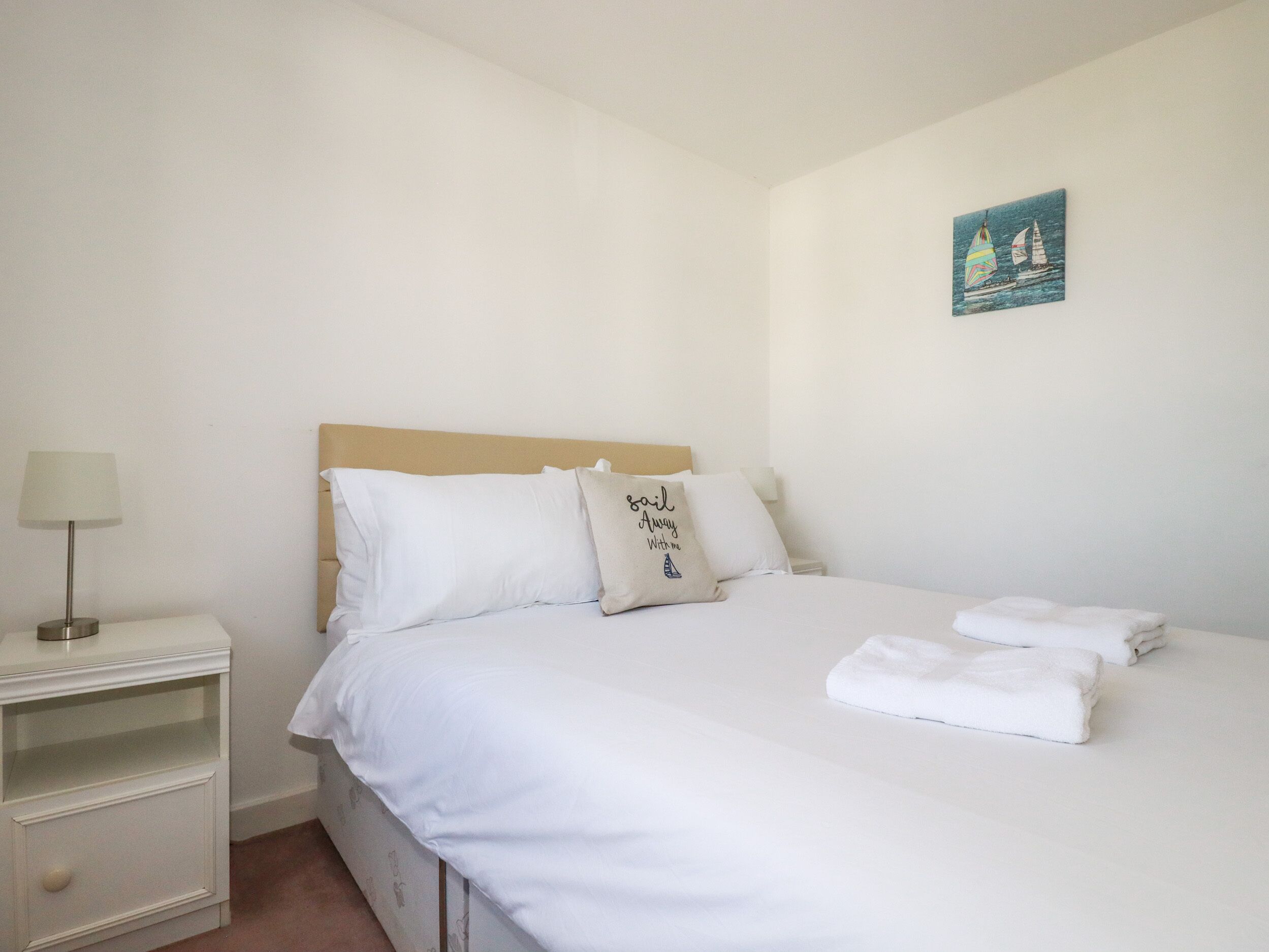 Una cama bien hecha con sábanas blancas, un cabecero color beige y una almohada con texto. Hay una mesita de noche pequeña con una lámpara y un cuadro colgado en la pared que muestra veleros.