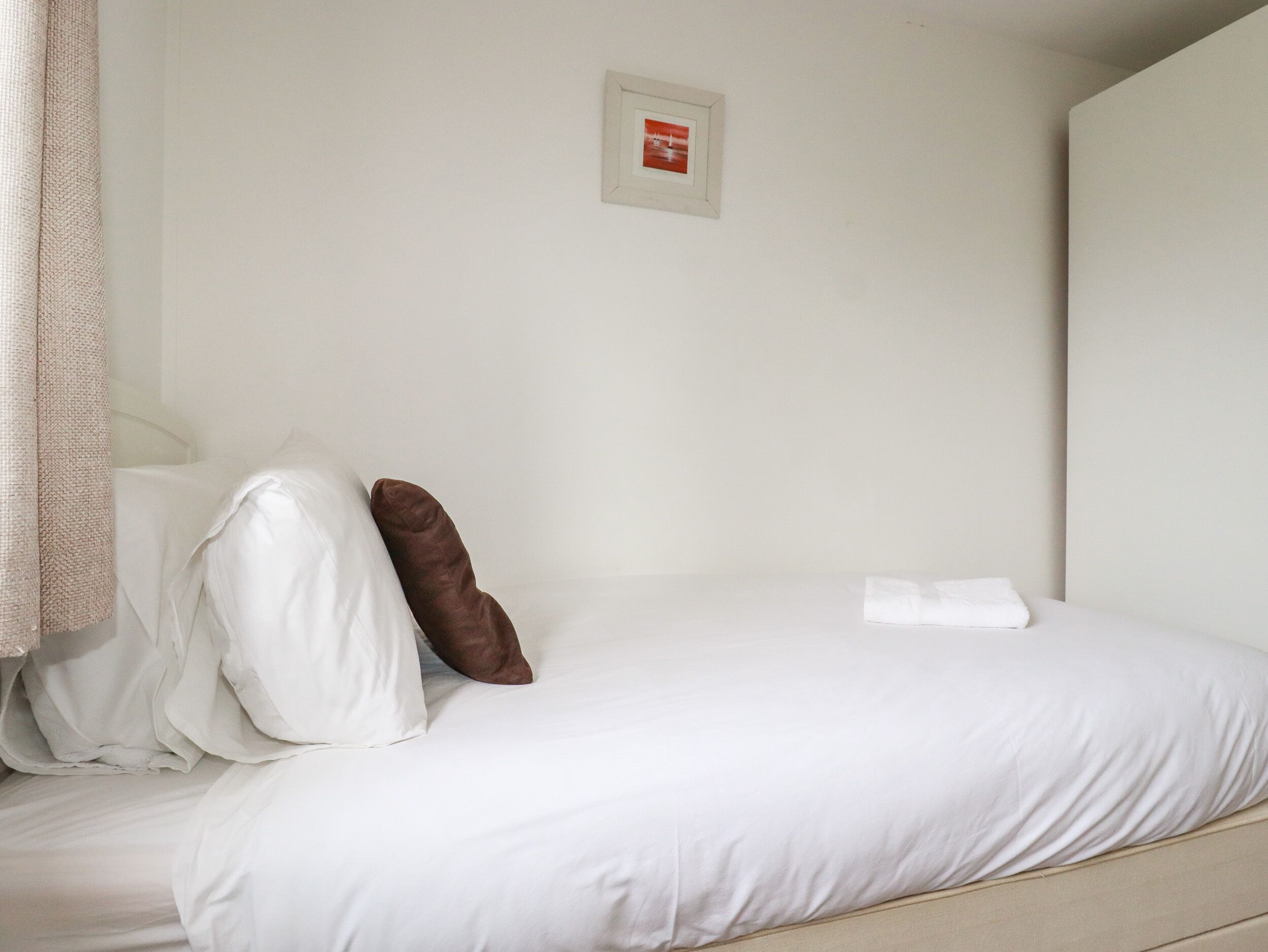 Una cama bien hecha con ropa de cama blanca y una almohada marrón, un cuadro en la pared y una toalla blanca sobre la cama.