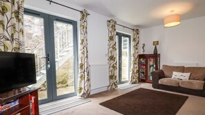 A living room with a TV, sofa, and a view of a stone building through the glass door. - Spinnaker (St Austell)