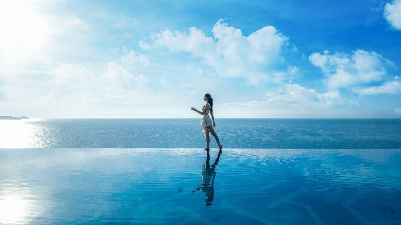 A person standing on a reflective infinity pool with a clear view of the ocean and sky.