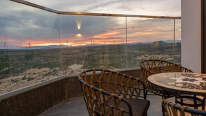 A corner room with a glass wall overlooking a desert landscape with wind turbines, furnished with wicker chairs and a round table.