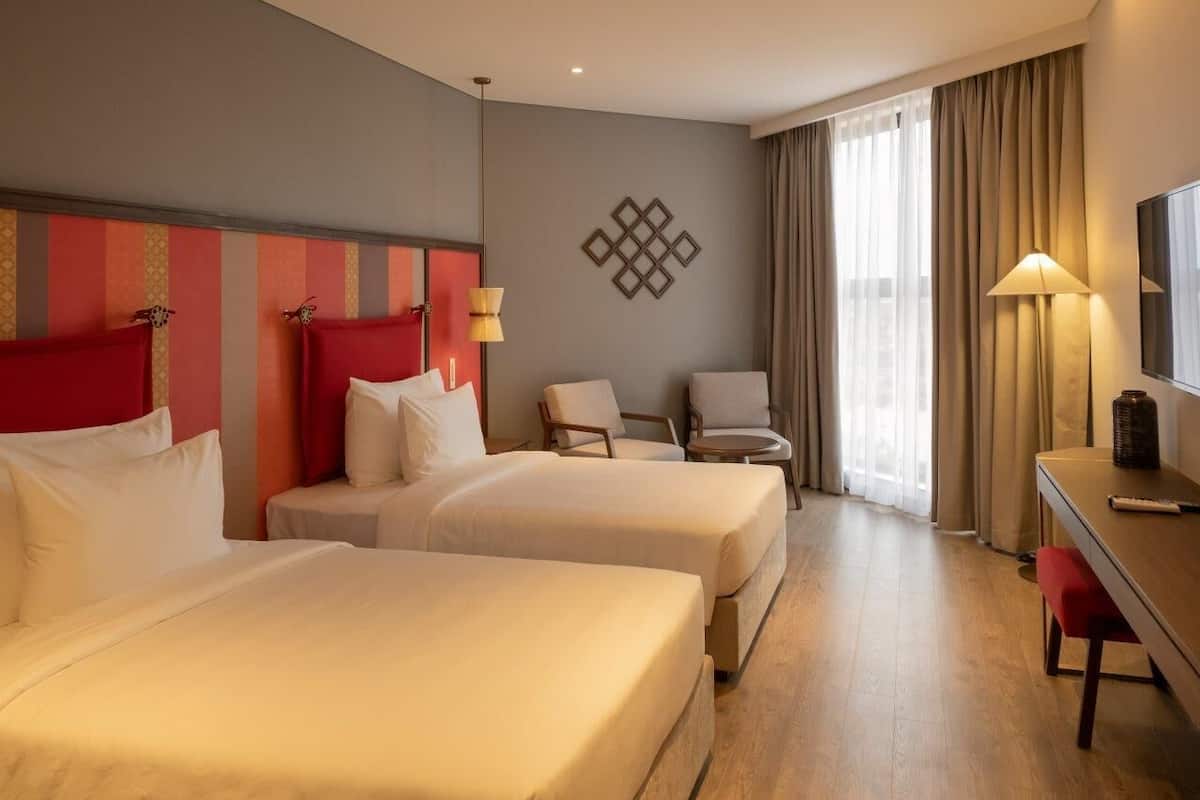 Een hotelkamer met twee bedden, een bureau, een stoel, een lamp en een groot raam met gordijnen.