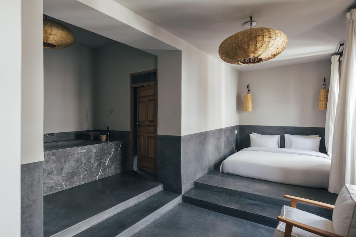 Un dormitorio moderno con cama, una silla y un baño con lavamanos y espejo.