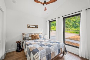 A bedroom with a large bed, a ceiling fan, a window with a view of greenery, and a small table with a candle.