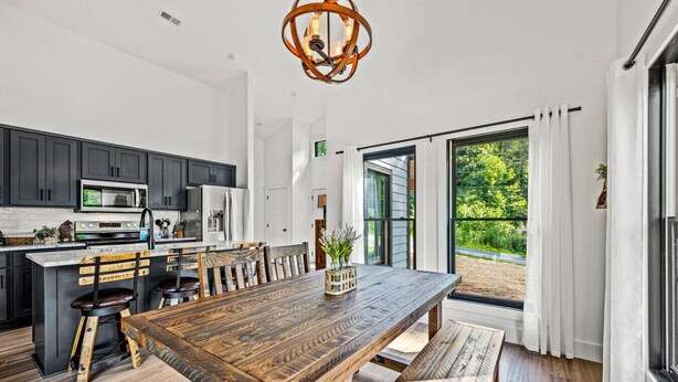 A modern kitchen with dark cabinets, a wooden dining table, and a large window offering a view of greenery.