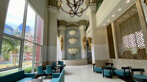 A spacious lobby with a high ceiling, a chandelier, and a view of palm trees and buildings outside. - Grande Caribbean by PSR (Pattaya)