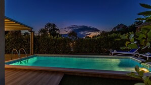 A swimming pool with a wooden deck, surrounded by a hedge and a house in the background. - Antonela in Malinska (Malinska-Dubasnica)