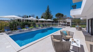 A modern outdoor pool area with lounge chairs and umbrellas, surrounded by greenery and a clear blue sky. - Frani in Pore With 4 Bedrooms and 4 Bathrooms (Porec)