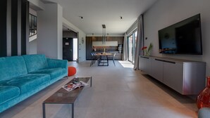 Een moderne woonkamer met een blauwgroene bank, een salontafel met een tijdschrift, een keuken op de achtergrond en een grote tv aan de muur.