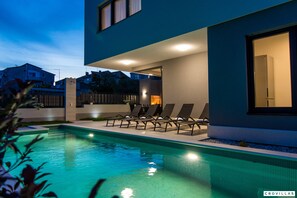 Ein modernes Gebäude mit Schwimmbad, Liegestühlen und einer gut beleuchteten Fassade.