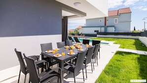 An outdoor dining area with a table set for breakfast, surrounded by chairs, and a view of a swimming pool and buildings in the background. - Kastel Two in Ka tel Kambelovac (Kastela)