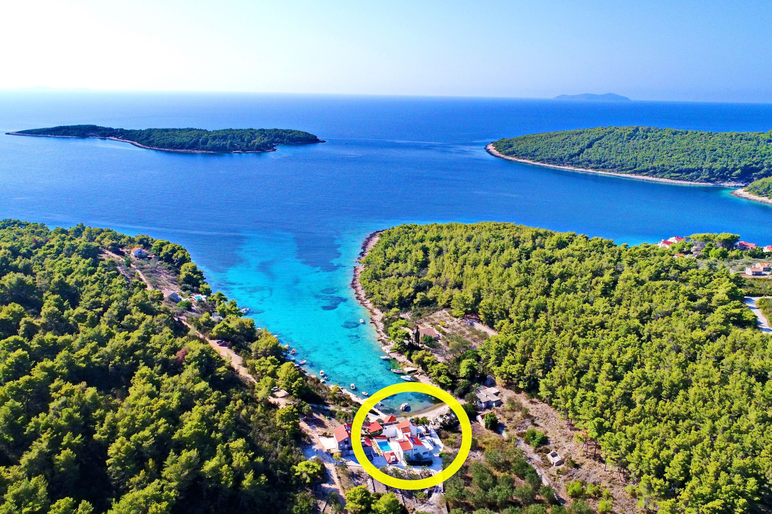 Vista aérea de una zona costera con un pequeño grupo de edificios rodeados de un bosque verde y frondoso, cerca de un mar azul y transparente.
