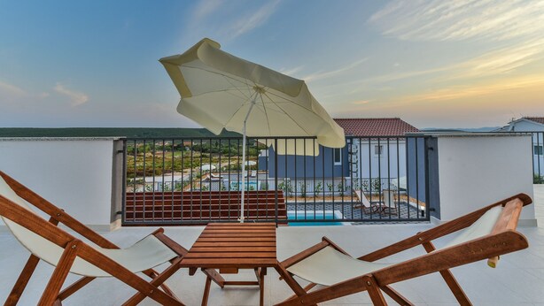 Un balcón con mesa y sillas de madera, un paraguas grande y vistas a una piscina y edificios al fondo.