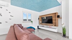 A modern living room with a large flat-screen TV mounted on a wooden wall, a pink sectional sofa, a small side table, and a potted plant. - Casa Mara in Tokovci (Svetvincenat)